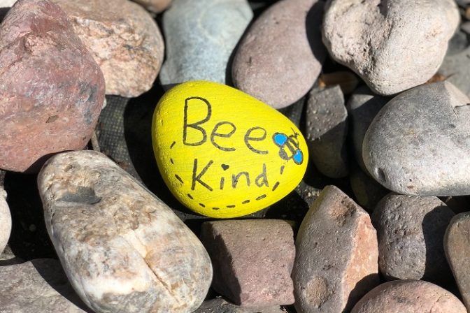 A yellow pebble lying among lots of other pebbles, with the words Be Kind written on it.